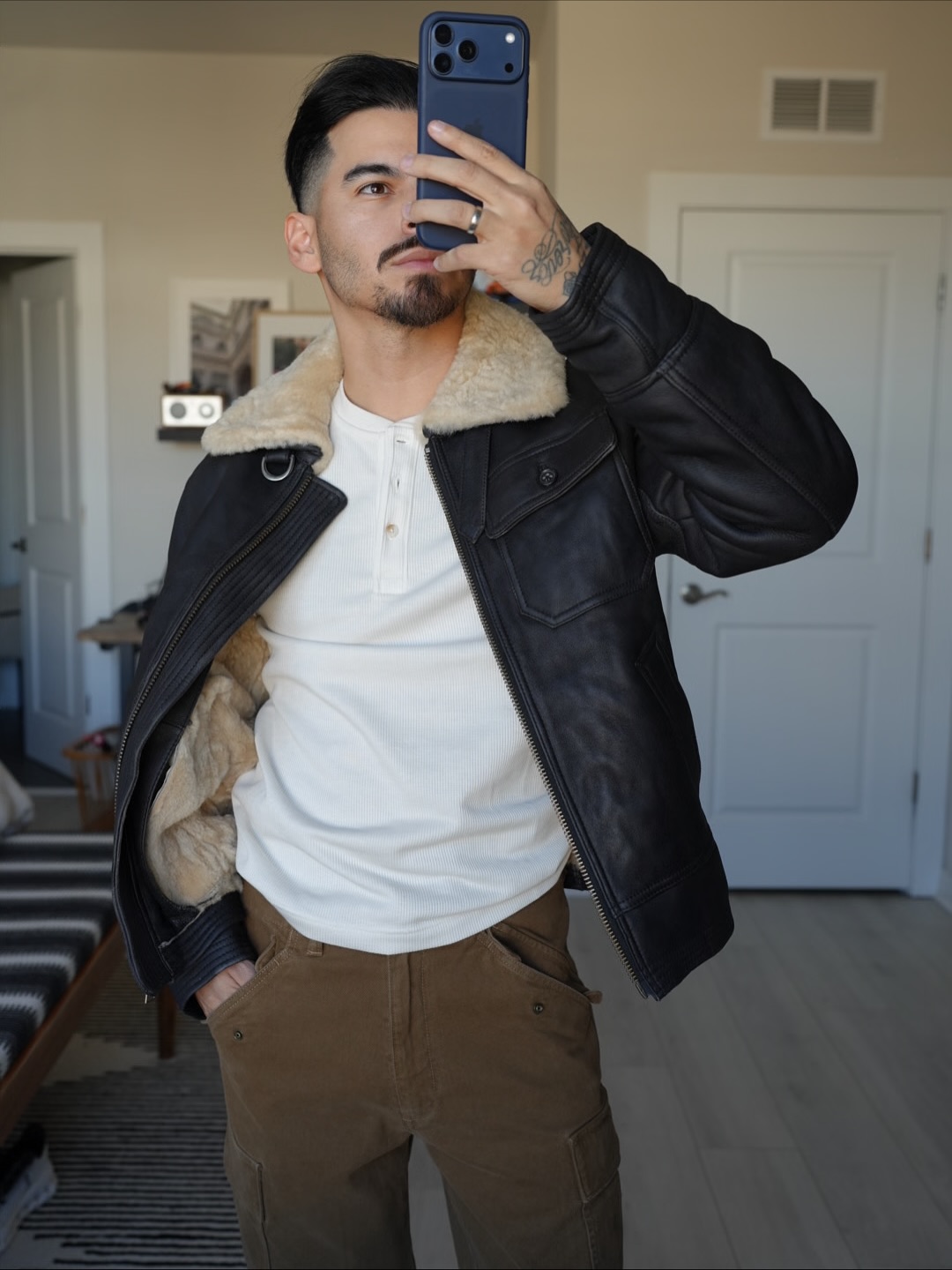 Rob in a black leather shearling jacket, white henley, and brown cargo pants